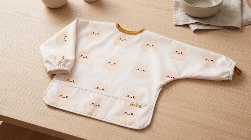 HAPSI Ärmellätzchen Klecksi Edition mit Klecksi-Motiven auf einem Holztisch, daneben Schüsseln und ein Leinentuch